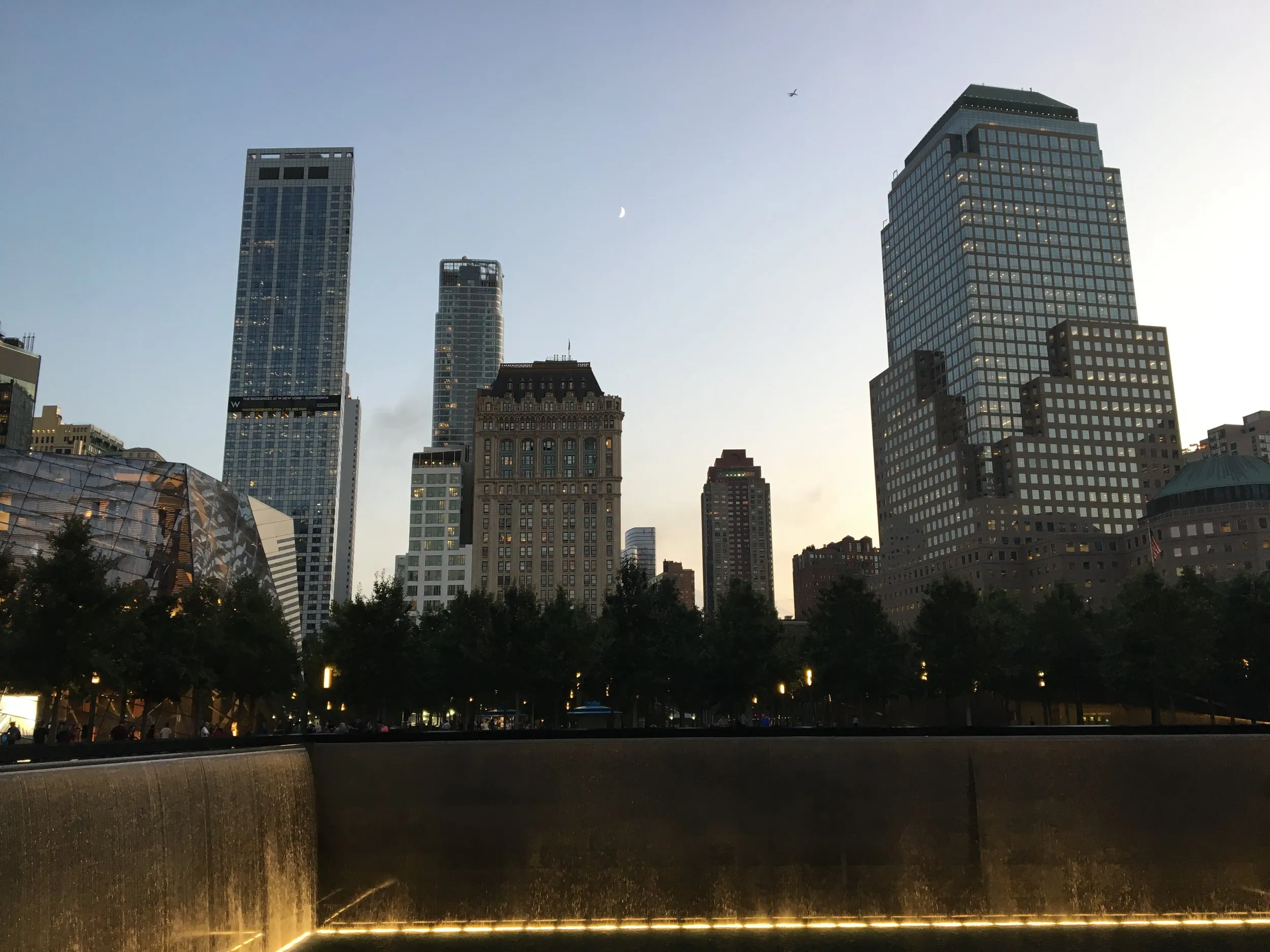 Glass towers at dusk, a moon, a plane, and the water catching all of it.
